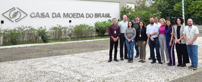 grupo de pessoas posam para foto em frente a parede que contém a inscrição Casa da Moeda do Brasil ao lado da logomarca da empresa