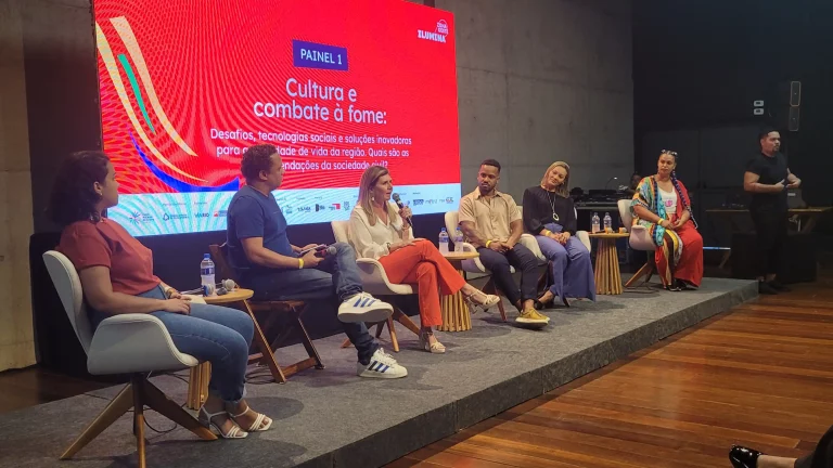 foto mostra palestrantes sentados em um palco raso e com telão ao fundo